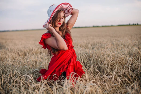 Kırmızı elbiseli ve çizgili şapkalı güzel kız buğday tarlasında poz veriyor. — Stok fotoğraf