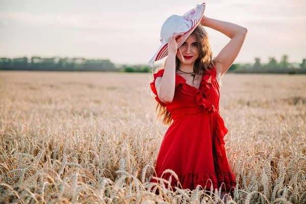 Vacker flicka i röd klänning och randig hatt poserar i ett vetefält — Stockfoto