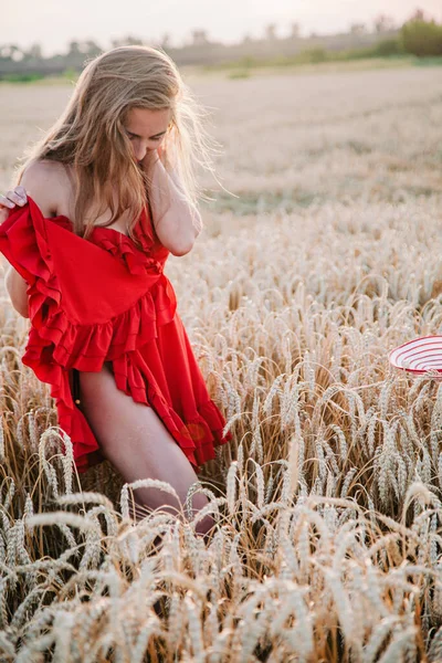 Vacker flicka i röd klänning och randig hatt poserar i ett vetefält — Stockfoto