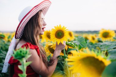 Kırmızı elbiseli, ince, genç bir kız ayçiçeği tarlasında poz veriyor.