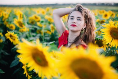 Kırmızı elbiseli, ince, genç bir kız ayçiçeği tarlasında poz veriyor.