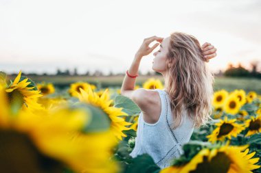 Beyaz tişörtlü genç, narin kız güneş batarken ayçiçeği tarlasında poz veriyor.