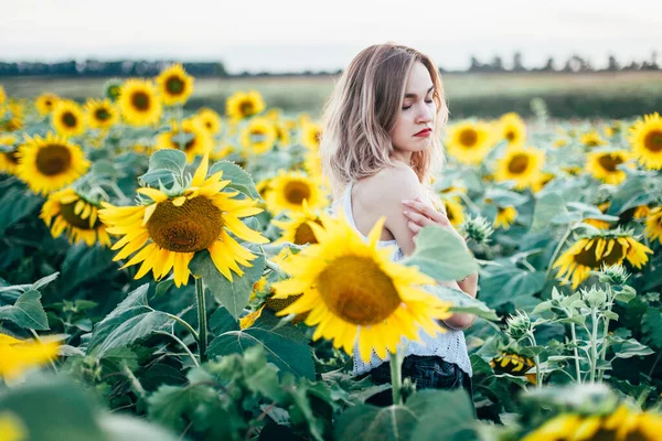 Mladá štíhlá dívka v bílém tričku pózuje při západu slunce na poli slunečnic — Stock fotografie