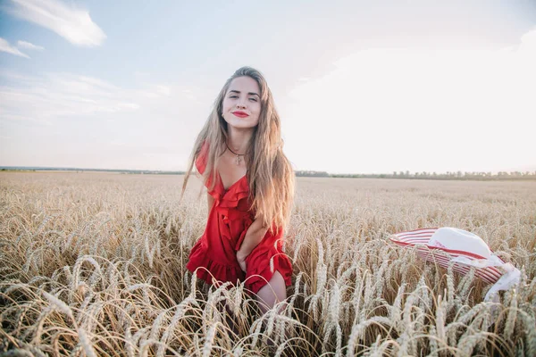 Kırmızı elbiseli ve çizgili şapkalı güzel kız buğday tarlasında poz veriyor. — Stok fotoğraf