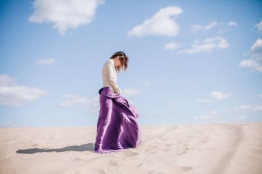 Çölde rüzgarda poz veren bej elbiseli, mor elbiseli genç bir kız.