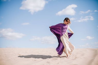 Çölde rüzgarda poz veren bej elbiseli, mor elbiseli genç bir kız.