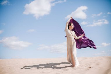 Çölde rüzgarda poz veren bej elbiseli, mor elbiseli genç bir kız.