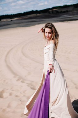 Çölde rüzgarda poz veren bej elbiseli, mor elbiseli genç bir kız.