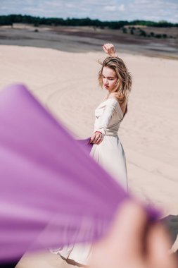 Çölde rüzgarda poz veren bej elbiseli, mor elbiseli genç bir kız.