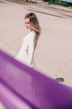 Çölde rüzgarda poz veren bej elbiseli, mor elbiseli genç bir kız.