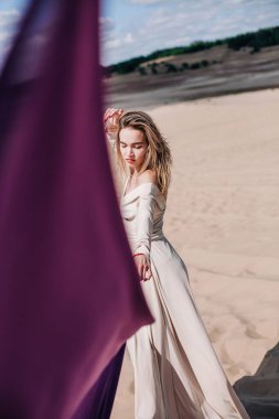 Çölde rüzgarda poz veren bej elbiseli, mor elbiseli genç bir kız.