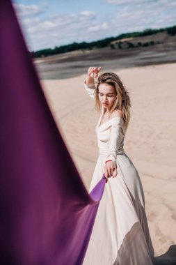 Çölde rüzgarda poz veren bej elbiseli, mor elbiseli genç bir kız.