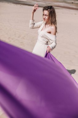 Çölde rüzgarda poz veren bej elbiseli, mor elbiseli genç bir kız.