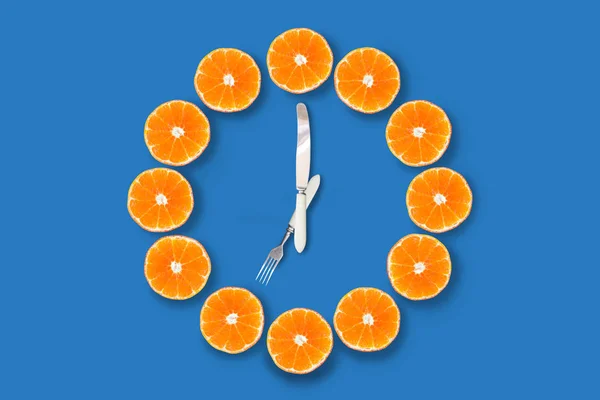 青い背景に7時を示すフォークとナイフで作られた矢印でハーフカットのみかんで作られた時計 良い健康な朝の概念 — ストック写真