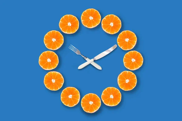 半分カットされたみかんで作られた時計は 青い背景に10分を過ぎて10分を示すフォークとナイフで作られた矢印で作られました 良い健康な朝の概念 — ストック写真
