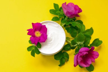 Parlak sarı arka planda, köpekli güllerin açtığı bir kavanozda beyaz krema. Çiçekler, üst görünüm. Yaşlanma önleyici krem, köpek yağı ve E vitamini.