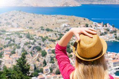 Güneş gözlüklü dalgalı saçlı genç kız dağın tepesinde oturuyor ve Dodekanisos takımadalarındaki Yunan adası Symi 'deki küçük renkli evleri ve Akdeniz' i izliyor.