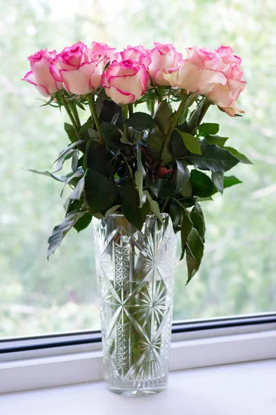 Freshly cut pink roses with dark green leaves in a cut-glass vase on white windowsill. Vertical format.