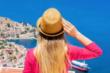 Hasır şapkalı, pembe uzun kollu, dalgalı saçlı genç bir kız dağın tepesinde dikiliyor ve Dodekanisos takımadasındaki Yunan adası Symi 'deki küçük renkli evleri ve Akdeniz' i izliyor. 
