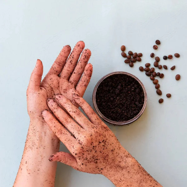 Top view of female hands and a jar with roasted coffee bean scrub and sea salt on mint background in square format. Rejuvenating scrub in round jar for skin exfoliation.