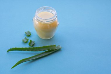 Krem ve aloe esanslı şeffaf cam kavanoz. Aloe bitkisi dilimleriyle mavi zemin üzerinde. Yumuşak kremin organik nemlendirici olarak canlandırılması.