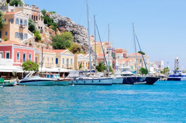 Kayalıklardaki renkli evlerin ve Yunan adası Symi 'deki derin mavi Akdeniz kıyısındaki yelkenli teknelerin manzarası güneşli yaz günlerinde, egzotik adalarda tatiller.