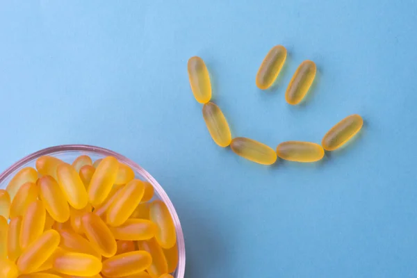 A lot of soft jelly capsules with fish oil as a source of Omega 3 in a transparent  bowl on blue background. Smile made of pills filled by fish oil. Concept of health care and using biologically active additives.