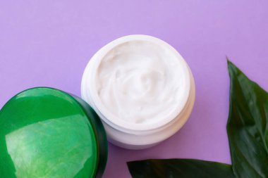 Kavanozda yumuşak beyaz krema ve mor arka planda parlak yeşil yapraklar. Evde spa tedavisi ve prosedür.