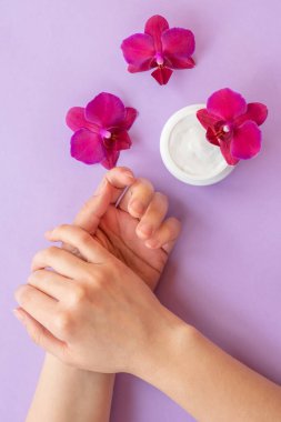 Yumuşak beyaz krem ve parlak mor orkideli dikey eller. Kavanozda canlandırıcı yüz kremi ve mor arka planda çiçek açan orkide, cilt bakımı kozmetiği..