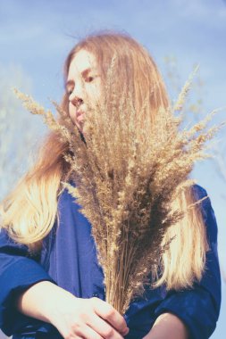 Güzel sarı saçlı, parlak güneş ışığında genç bir kadın, açık mavi gökyüzüne karşı, bir buket kurutulmuş sarı otla.