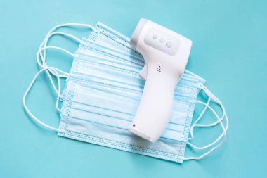 Dijital kızılötesi termometre mavi arka plandaki tıbbi maske yığınında yalan söylüyor. Virüs ve gribe karşı sağlık koruma araçları 