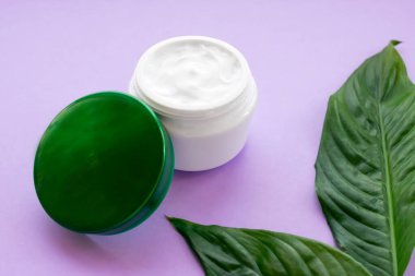 Kavanozda yumuşak beyaz krema ve mor arka planda parlak yeşil yapraklar. Evde spa tedavisi ve prosedür.