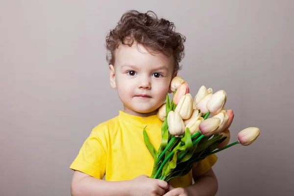 Little boy in yellow shirt gives a bouquet of tulips on light background