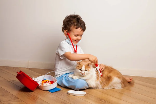 funny curly kid in a medical mask and glasses with a stethoscope on his neck plays a doctor with a cat. The child wants to inject the cat with a toy syringe.