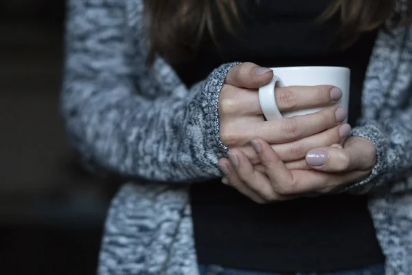 The girl warms her hands on a white cup of warm tea