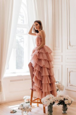 Pahalı parfüm reklamı tarzında bir fotoğraf. Eski bir stüdyoda küçük bir merdivende duran bir kız penceresinin yanında taze şakayıklar, tek bacağı açık ve güzel ve şık pembe bir elbise giyiyor.