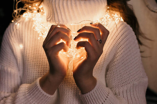 Девушка в белом свитере, завернутая в рождественскую гирлянду