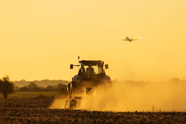 现代拖拉机犁在夕阳与大量的灰尘背景和飞机起飞 — 图库照片