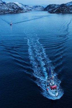 Balıkçı teknesi güneş doğmadan önce arka planda karla dolu dağlarla balığa çıkıyor. Lofoten Adası, Norveç.