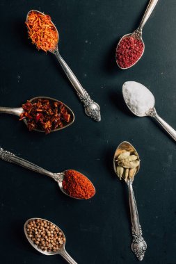 top view of different spices in spoons on dark table clipart