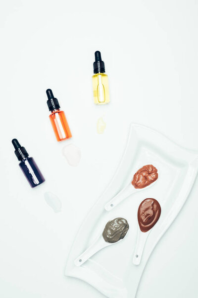 top view of clay powder, cosmetic bottles with pipes, plate and spoons with various colorful clay masks isolated on white surface 