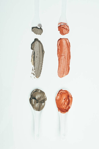 flat lay with spoons and various colorful clay mask smudges placed in row isolated on white surface 