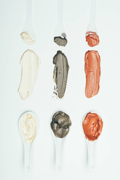 flat lay with spoons and various colorful clay mask smudges placed in row isolated on white surface 