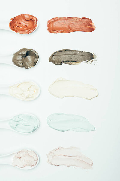 top view of spoons and various colorful clay mask smudges placed in row isolated on white surface 