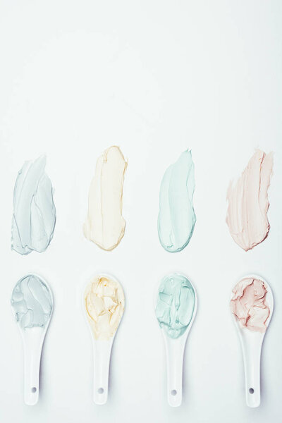 top view of spoons and colorful clay mask smudges placed in row on white surface 