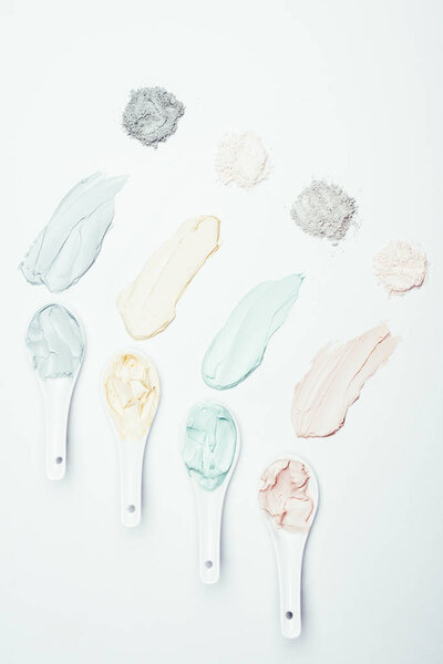 flat lay with spoons, different clay powder and colorful clay mask smudges placed in row on white surface 