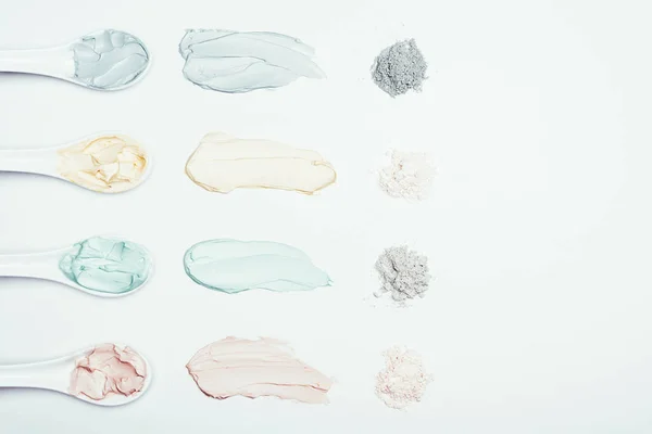 flat lay with spoons, clay powder and colorful clay mask smudges placed in row on white surface