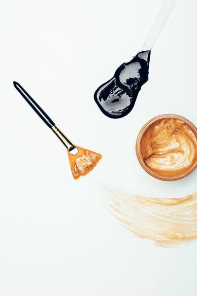 верхний вид золотой маски в контейнере, кисти, пятна и ложки с черной маской изолированы на белой поверхности
 