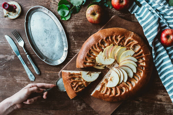 partial view of woman with cake server taking piece of homemade apple pie on wooden cutting board
