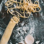 Top view of dough, egg shell and raw pasta on table covered by flour